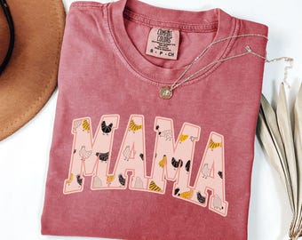 Op de afbeelding: Een rood T-shirt met het woord "MAMA" erop gedrukt in roze met een witte omtrek. De letters zijn versierd met kleine zwarte en gele kippen. Het shirt heeft een verweerde look.