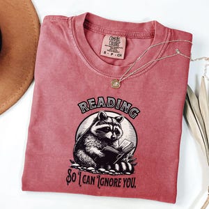 May include: A red t-shirt with a graphic of a raccoon reading a book. The text on the shirt reads "Reading so I can ignore you."