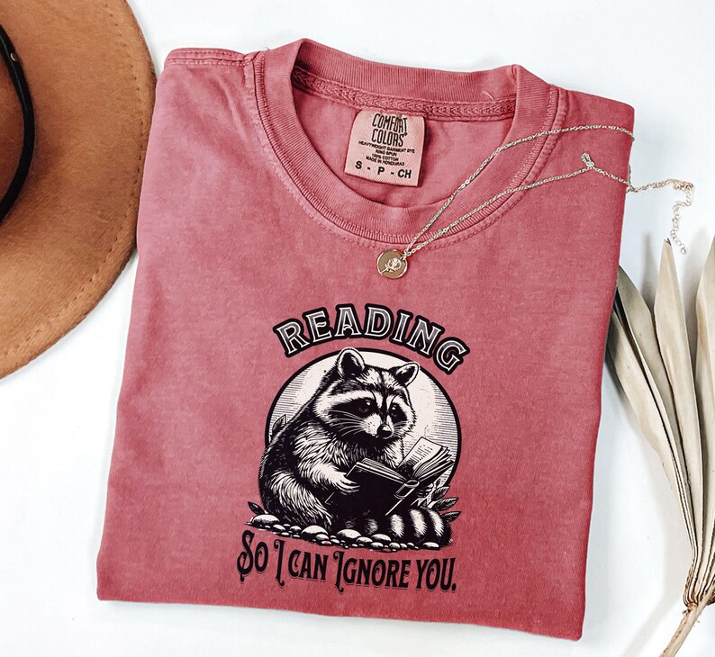May include: A red t-shirt with a graphic of a raccoon reading a book. The text on the shirt reads "Reading so I can ignore you."