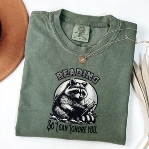 May include: A green t-shirt with a black and white illustration of a raccoon reading a book. The text on the shirt reads "Reading so I can ignore you."