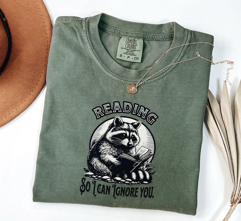 May include: A green t-shirt with a black and white illustration of a raccoon reading a book. The text on the shirt reads "Reading so I can ignore you."