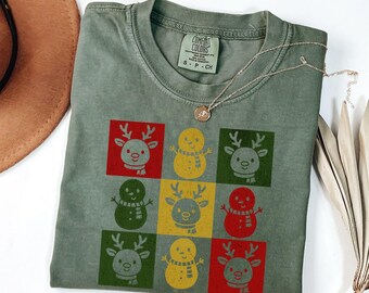 May include: Green Comfort Colors t-shirt featuring a grid of Christmas-themed graphics. The design includes reindeer and snowman characters in red, yellow, and green squares. A silver necklace with a pendant is draped across the shirt.