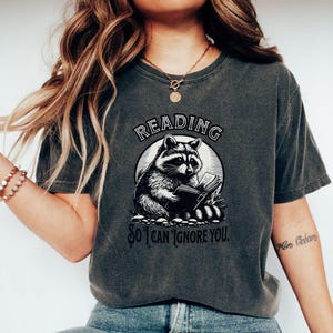 May include: A dark gray t-shirt with a graphic of a raccoon reading a book. The text on the shirt reads "Reading so I can ignore you."