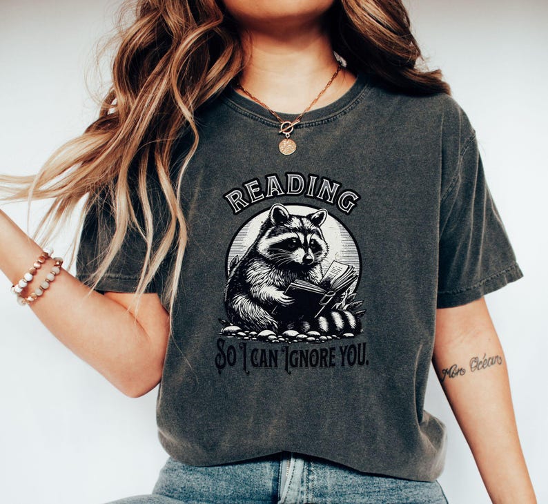 May include: A dark gray t-shirt with a graphic of a raccoon reading a book. The text on the shirt reads "Reading so I can ignore you."