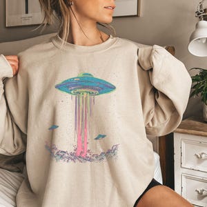 May include: A beige sweatshirt featuring a graphic print of a flying saucer with rainbow beams of light emanating from it. Smaller flying saucers surround the main graphic.