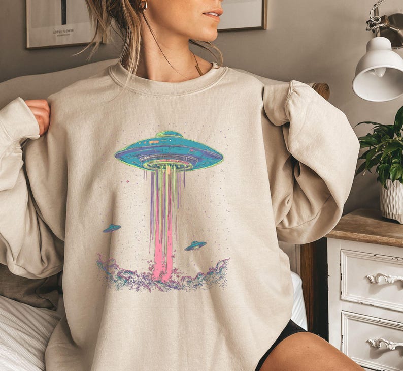 May include: A beige sweatshirt featuring a graphic print of a flying saucer with rainbow beams of light emanating from it. Smaller flying saucers surround the main graphic.