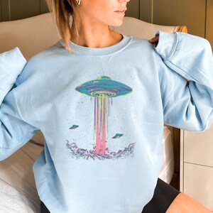 May include: A light blue sweatshirt featuring a graphic of a flying saucer with rainbow beams of light emanating from it. The saucer is surrounded by stars and there are two smaller flying saucers in the background.