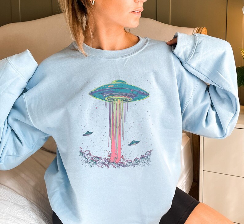 May include: A light blue sweatshirt featuring a graphic of a flying saucer with rainbow beams of light emanating from it. The saucer is surrounded by stars and there are two smaller flying saucers in the background.