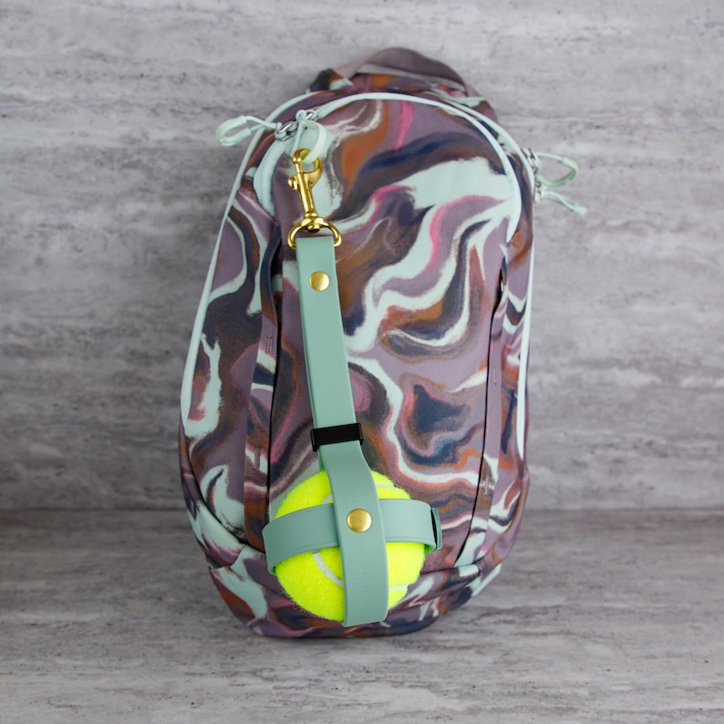 May include: A backpack with a swirling pattern in shades of purple, brown, and pink. A light green strap with a gold-colored clasp holds a yellow tennis ball. The strap is attached to the backpack.