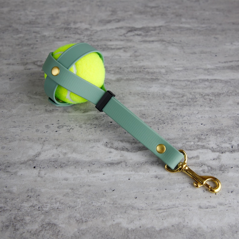 May include: A dog toy featuring a bright yellow tennis ball held by a seafoam green strap with gold-colored hardware. The strap has a black accent and a gold-colored clasp.