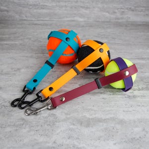 May include: Three dog toy tennis balls with colorful straps and black or silver clips. The balls are orange, black, and yellow-green. The straps are teal, orange, and burgundy. The clips are black and silver.