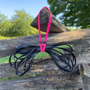 May include: A long black dog leash featuring a bright neon pink handle loop and a matching pink connector strap. The leash is made of a smooth, flat material and includes a black metal swivel snap hook at the end for secure attachment.