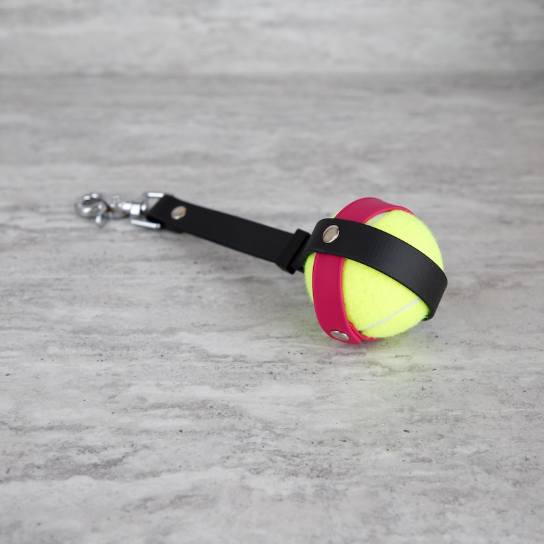 May include: A tennis ball keychain with a black leather strap and a silver-tone clasp. The tennis ball is secured by a pink leather band. The keychain is designed for sports enthusiasts and pet owners.