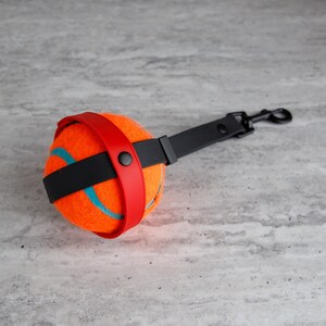 May include: An orange tennis ball with blue curved lines, secured by a red and black strap, and a black handle with a metal clip. The dog toy is designed for interactive play.