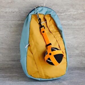 May include: A mustard yellow and light blue backpack with a black and orange ball holder attached. The ball holder has an orange strap that wraps around a black and white ball. The backpack has a zipper and a silver clasp.