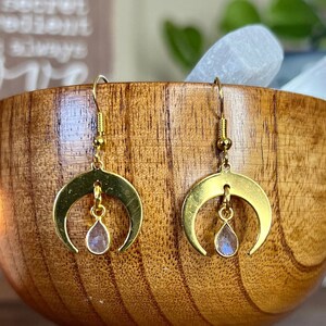 May include: Gold crescent moon earrings with a teardrop-shaped iridescent gemstone hanging from each moon. The earrings are displayed in a wooden bowl.