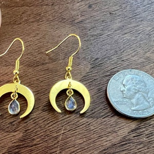 May include: A pair of gold crescent moon earrings with a teardrop-shaped clear crystal hanging from each moon. The earrings are shown next to a UK 20 pence coin for size comparison.