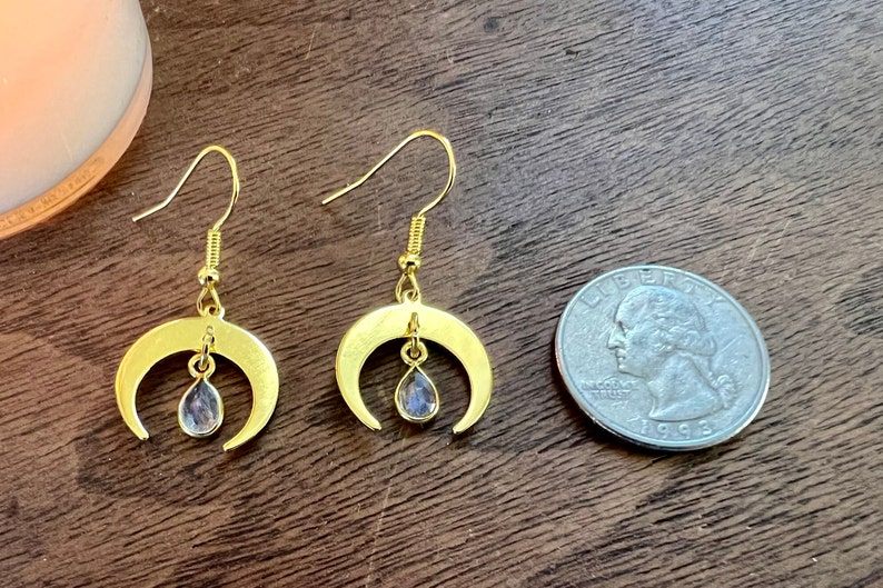 May include: A pair of gold crescent moon earrings with a teardrop-shaped clear crystal hanging from each moon. The earrings are shown next to a UK 20 pence coin for size comparison.