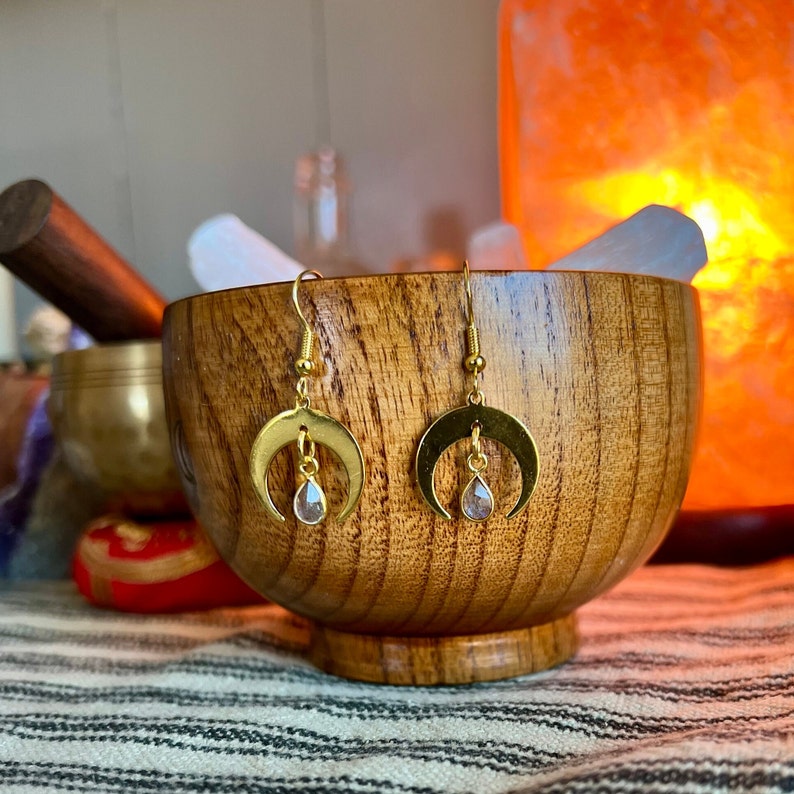 May include: A pair of gold crescent moon earrings with a teardrop-shaped clear crystal hanging from each moon. The earrings are displayed in a wooden bowl.