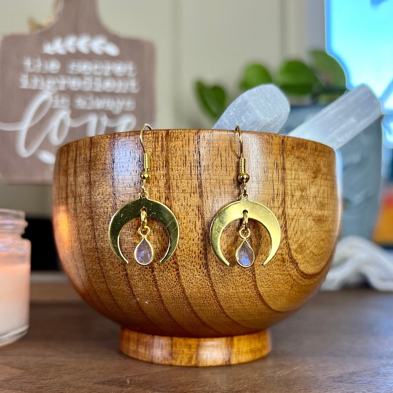 May include: Gold crescent moon earrings with a teardrop-shaped iridescent gemstone hanging from each moon. The earrings are displayed in a wooden bowl.