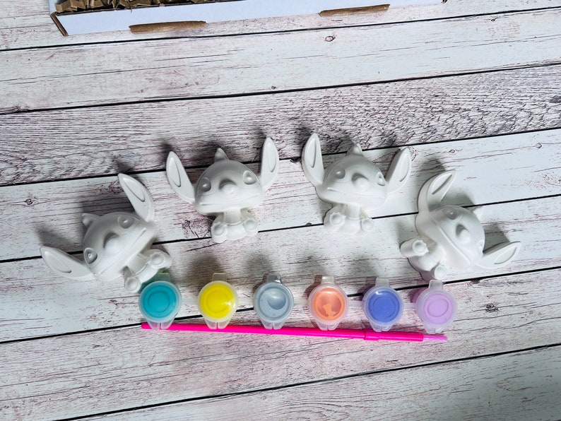 May include: Four white ceramic figurines of Stitch from Lilo & Stitch, ready to be painted. The set includes six small paint pots in blue, yellow, gray, orange, purple, and pink, and a pink paintbrush.