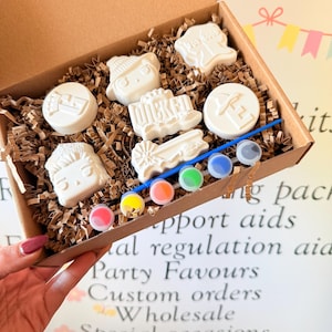 May include: A craft kit in a cardboard box, containing white plaster figures, paint pots, and a paintbrush. The figures include a hat, a face, and the word "Wicked". The kit is designed for creative activities.