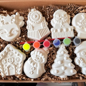 Puede incluir: Un kit de manualidades con ocho adornos navideños de cerámica sin pintar en una caja de cartón. Los adornos incluyen un reno, un tren, Papá Noel, un globo de nieve, una casa de jengibre, un muñeco de nieve, un árbol de Navidad y un hombre de jengibre. Se incluyen botes de pintura y pinceles.