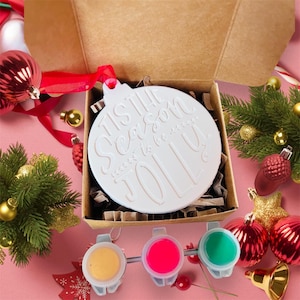 May include: A white ceramic Christmas bauble in the shape of a ball with the text "Tis the Season to be Jolly" in a white script font. The bauble is in a brown box with white shredded paper. Three small paint pots with yellow, pink, and green paint are in front of the box.