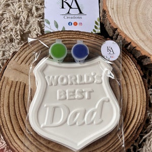 May include: White ceramic plaque shaped like a shield with the words "World's Best Dad" in raised lettering. The plaque is packaged with two small pots of paint, one green and one blue.