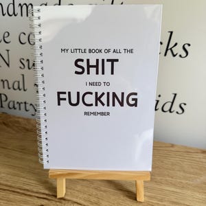 May include: A white spiral-bound notebook with black text on the cover that reads: MY LITTLE BOOK OF ALL THE SHIT I NEED TO FUCKING REMEMBER. The notebook is displayed on a small, light-coloured wooden easel stand.