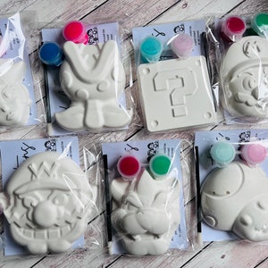 May include: Set of six white ceramic paint-your-own figurines with paint pots and brushes. The figurines include a question mark block, a mushroom, a character with a mustache, a character with a crown, a character with a hat, and a character with a crown.