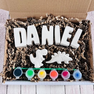 Puede incluir: Un kit de manualidades con letras de cerámica blanca que deletrean "DANIEL", dos figuras de dinosaurios y seis botes de pintura en negro, verde, amarillo, naranja, rojo y azul.