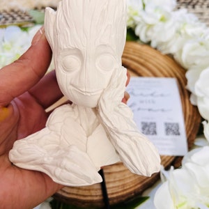 May include: White ceramic planter shaped like the character Groot from the Guardians of the Galaxy film. The planter has a detailed face and a textured surface.