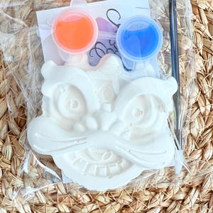 May include: A white plaster lion head craft kit in a clear plastic bag. The kit includes orange and blue paint pots, and a black paintbrush. The lion head has detailed features and is ready to be painted.