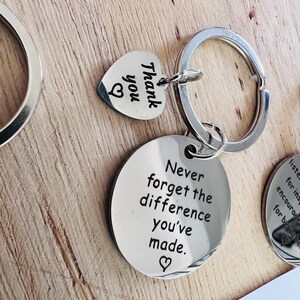 May include: Silver keyring with two charms. The larger charm is round and says "Never forget the difference you've made." The smaller charm is heart-shaped and says "Thank you."