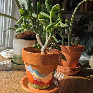 Peut inclure: Une plante succulente verte dans un pot en terre cuite avec une scène de montagne peinte à la main. Le pot a une base brun clair avec un bord vert et une soucoupe blanche.