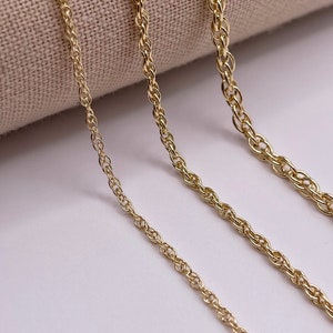 May include: Three gold rope chains of different lengths, laid out on a beige fabric background.
