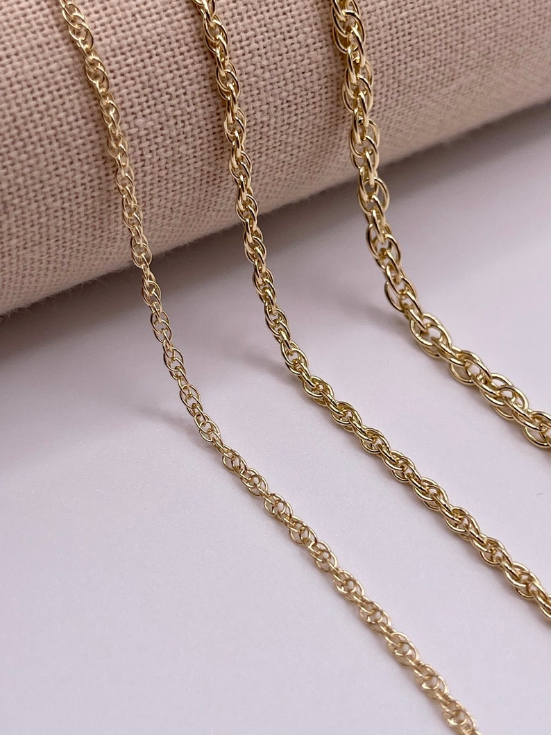 May include: Three gold rope chains of different lengths, laid out on a beige fabric background.