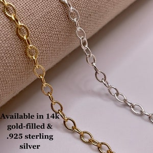 May include: Two chain necklaces, one gold-filled and one sterling silver, lay on a beige fabric surface. The text "Available in 14k gold-filled & .925 sterling silver" is visible below the chains.