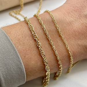 May include: Three gold chains of different thicknesses, laid out on a person's wrist. The chains are all made of a rope-like design.