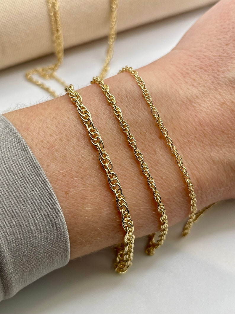 May include: Three gold chains of different thicknesses, laid out on a person's wrist. The chains are all made of a rope-like design.