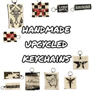 May include: A collection of handmade, upcycled keychains. Designs include a cross, stars, band logos, and graphic illustrations. Each keychain is attached to a metal ring. The keychains are made from fabric and feature various colors and patterns.