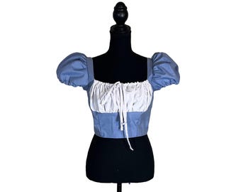 Puede incluir: Top corto azul claro con mangas cortas abullonadas y escote cuadrado. La parte superior presenta un corpiño blanco fruncido con un detalle de cordón. La prenda se exhibe en un maniquí negro sobre un fondo blanco.