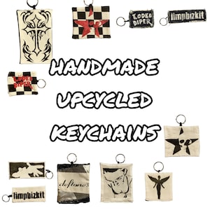 May include: A collection of handmade, upcycled keychains. The keychains feature various designs, including a cross, stars, and band logos. The keychains are made from fabric and have a metal ring for attaching keys.