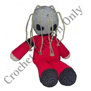 Puede incluir: Un juguete de peluche tejido a crochet en forma de figura humanoide con cabeza gris, cuerpo rojo y pies negros. El juguete tiene largos mechones de hilo gris que salen de su cabeza.