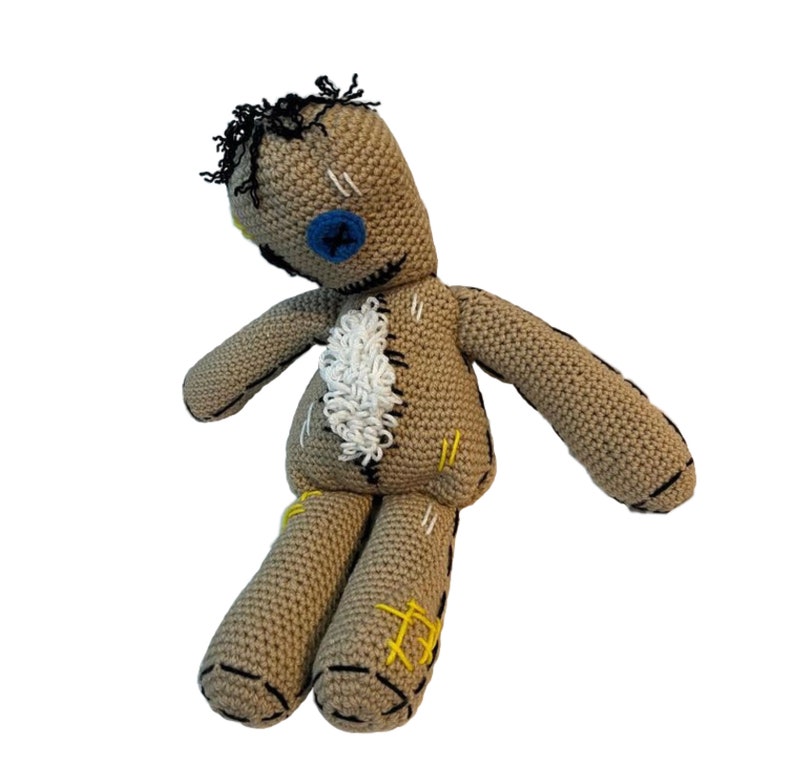 May include: A crocheted voodoo doll with a brown body, black hair, and a blue button eye. The doll has white and yellow stitching details.