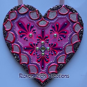 May include: A heart-shaped decorative piece with a pink ribbon for hanging. The heart features a central design in shades of pink and purple, embellished with glitter and rhinestones. The border is lined with dark glitter and the text "Raven Richl Creations" is at the bottom.