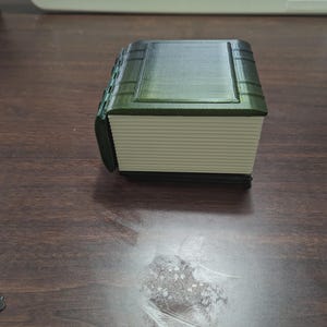 May include: A 3D-printed book-shaped box with a dark green cover and spine. The pages are a light cream color. The box is sitting on a dark brown wooden surface.