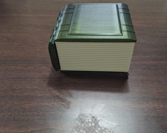 May include: A 3D-printed book-shaped box with a dark green cover and spine. The pages are a light cream color. The box is sitting on a dark brown wooden surface.