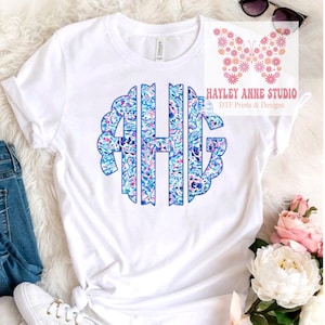 May include: White t-shirt with a blue monogram design featuring the letters "AHG" in a floral pattern. The monogram is surrounded by a blue circle with a scalloped edge. The shirt is folded and displayed on a white surface.